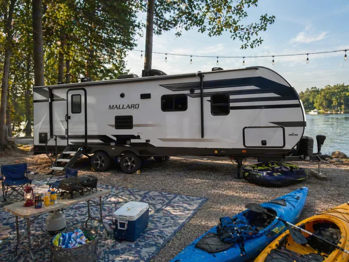 Lakeside RV rental: White travel trailer, kayaks, table, and chairs under string lights. Ideal outdoor adventure.. Heartland RVs Mallard 2023