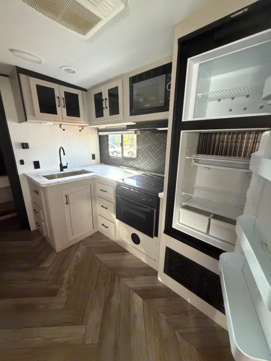 Bright RV rental kitchen with an L-shaped counter, sink, microwave, stove, and open Dometic refrigerator. Features modern cabinets and herringbone flooring.. Heartland RVs Mallard 2023