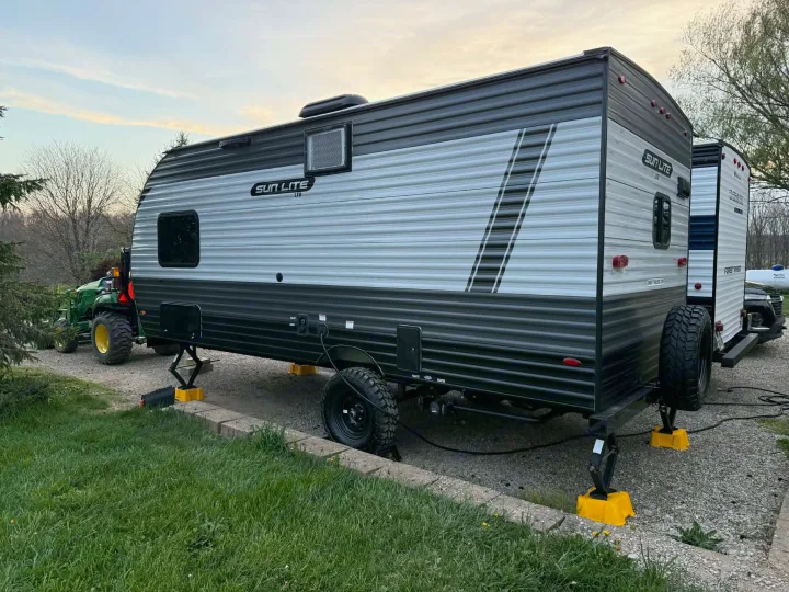 A grey and silver Sun Lite travel trailer rv rental, leveled on a gravel spot, awaits its next outdoor journey.. Sunset Park RV Sun Lite LTD 2025