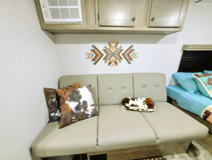 Cozy RV rental interior with a gray sofa, cowhide pillows, geometric wall art, and a comfortable bed with blue bedding.. Sunset Park RV Sun Lite LTD 2025