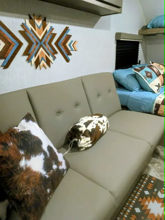 Inside an RV rental: a light grey sofa with cowhide & geometric pillows, Southwestern wall art, and blue bedding. Ready for adventure!. Sunset Park RV Sun Lite LTD 2025