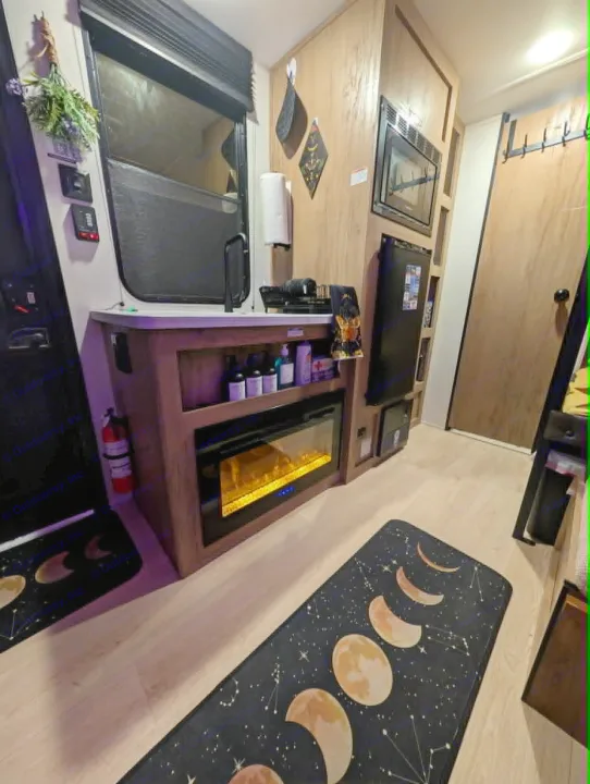 RV rental interior: kitchen with microwave & fridge, cozy electric fireplace, and a celestial moon phase rug on light wood flooring.. Forest River Cherokee Wolf Pup 2025