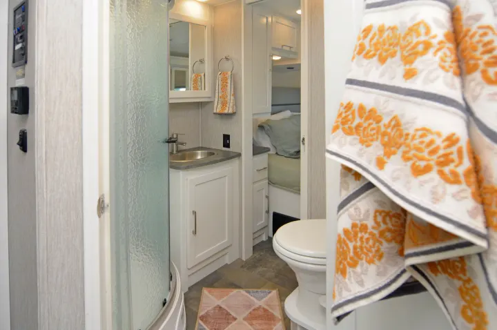 An RV rental bathroom features a compact shower, sink, and toilet, with a glimpse of the bedroom beyond. An orange patterned towel hangs.. Coachmen Pursuit 2022