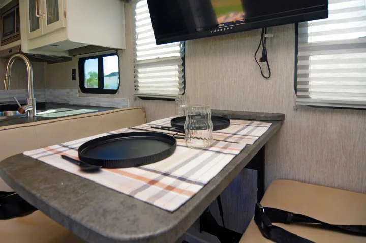 This RV rental's compact dinette and kitchen feature a table set with plaid placemats, plates, and glasses, a microwave, sink, and wall-mounted TV.. Coachmen Pursuit 2022