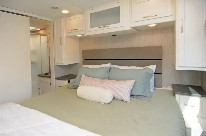 Modern RV rental bedroom features a comfy bed, overhead cabinets, and a sliding door to the bathroom with a sink and shower.. Coachmen Pursuit 2022