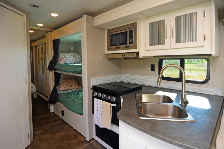 Modern rv rental interior showing a kitchen with double sink, stove, and microwave, next to two cozy bunk beds with curtains.. Coachmen Pursuit 2022