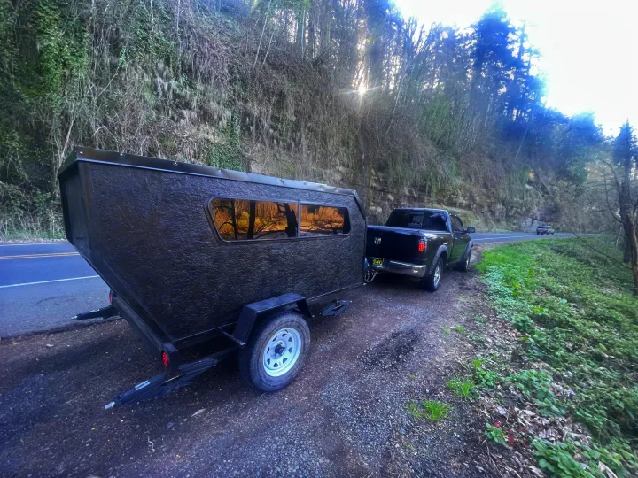Black truck pulling a textured camper trailer on a scenic road. This compact RV rental is ready for outdoor adventures.. Custom Squaredrop Trailer 2021