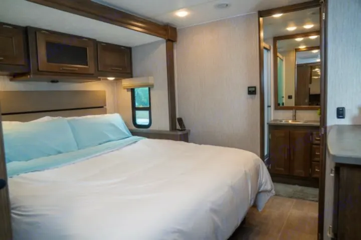 Cozy rv rental bedroom features a large bed with light blue sheets, overhead cabinets, and an adjacent bathroom vanity.. Forest River Coachmen Mirada 2022