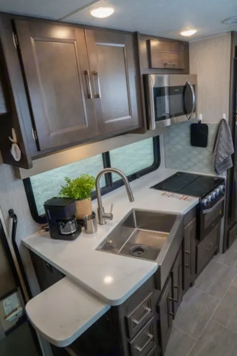 Modern rv rental kitchen: dark wood cabinets, white counters, stainless sink, microwave, stovetop, coffee maker, and pull-out extension.. Forest River Coachmen Mirada 2022