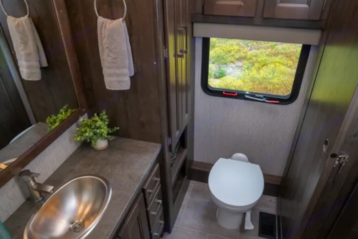 This modern RV rental bathroom features a stainless steel sink set in a vanity, a white toilet, mirror, and a window overlooking nature.. Forest River Coachmen Mirada 2022