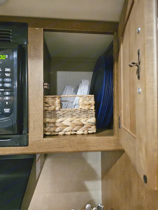Inside an RV rental kitchen cabinet: stacked plates, a woven basket, and microwave. Organized dish storage.. Grand Design Reflection 2016