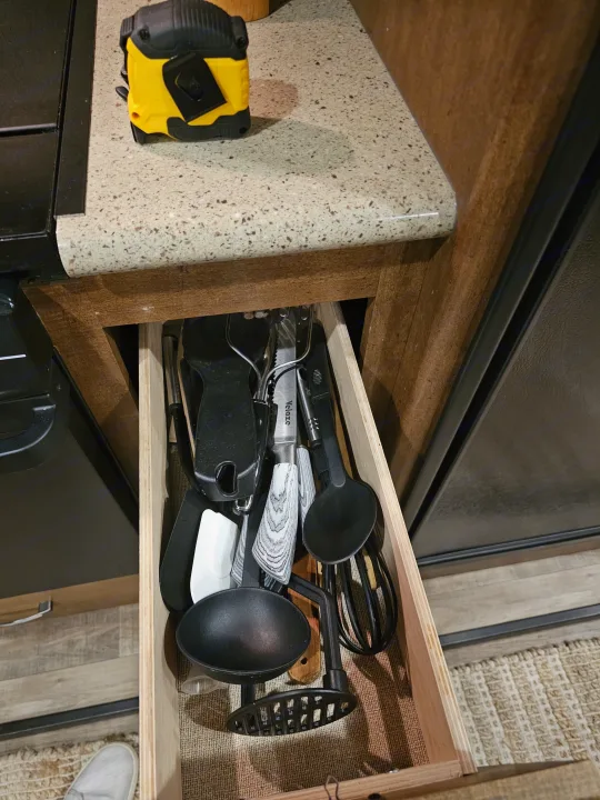 Open kitchen drawer in an RV rental, filled with various cooking utensils. A yellow tape measure sits on the speckled countertop.. Grand Design Reflection 2016