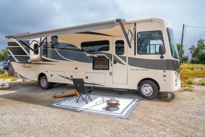 This RV rental offers a perfect outdoor setup: extended awning, rug, chair, fire pit, and cornhole boards on a gravel lot.. Forest River Coachmen Mirada 2022