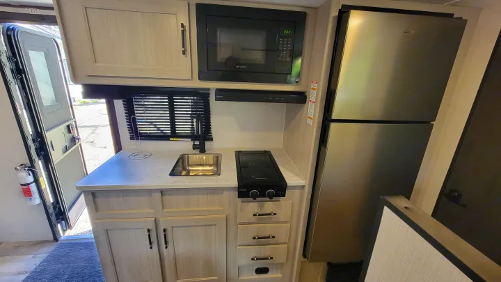 This RV rental kitchen features a microwave, Dometic fridge, Greystone stovetop, and sink, with an open door and fire extinguisher.. Crossroads RV Sunset Trail 2026