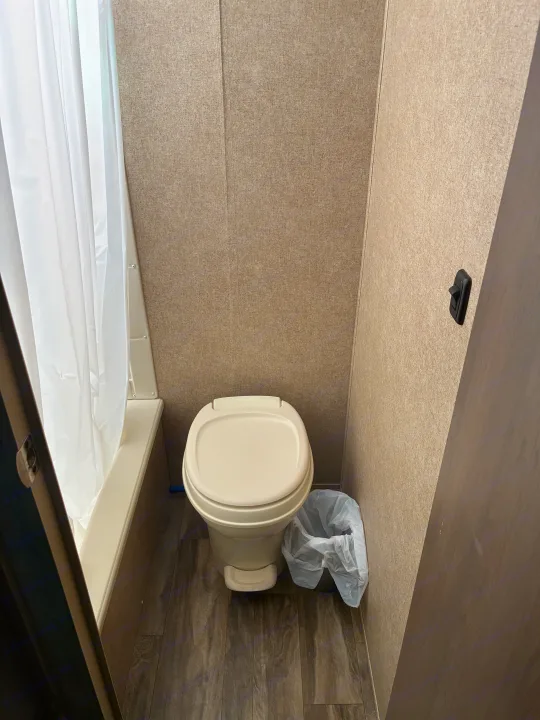 RV rental bathroom. Features a compact beige toilet, white shower curtain, and textured walls with wood-look floor.. Coachmen Catalina Sumit Series 2019