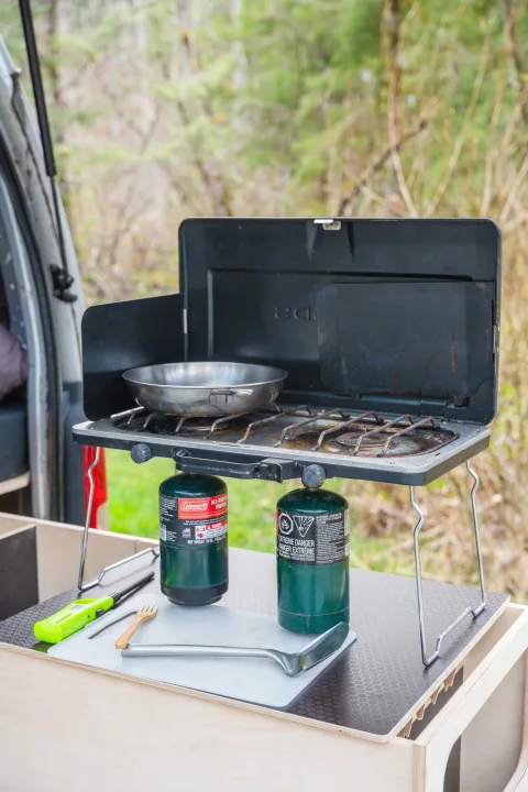RV rental: Portable Coleman propane camping stove with pan & utensils on a compact table for outdoor cooking.. Toyota Hiace 2005