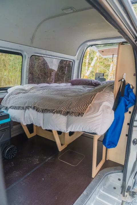 This camper van RV rental features a cozy bed with white bedding, a striped blanket, and a wooden frame overlooking nature.. Toyota Hiace 2005