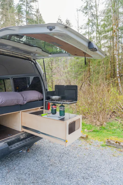 Back of an RV rental camper van with open tailgate, showing a pull-out kitchen, stove, and bed inside for camping.. Toyota Hiace 2005