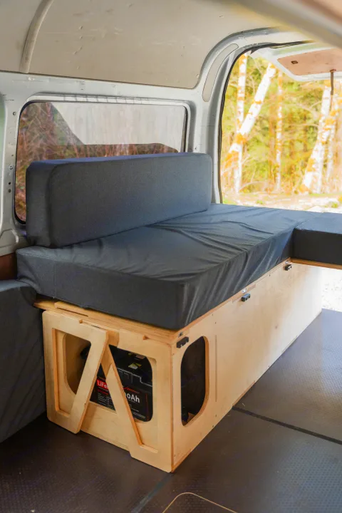 Inside an RV rental: a cozy van bed with storage, including a battery, and a window framing a forest view.. Toyota Hiace 2005