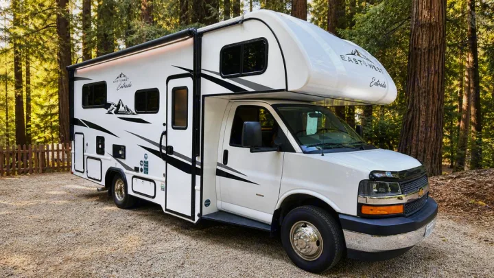 White East to West Entrada RV rental parked on gravel, surrounded by tall trees. Ideal for a nature-filled camping adventure.. East to West Entrada 2025