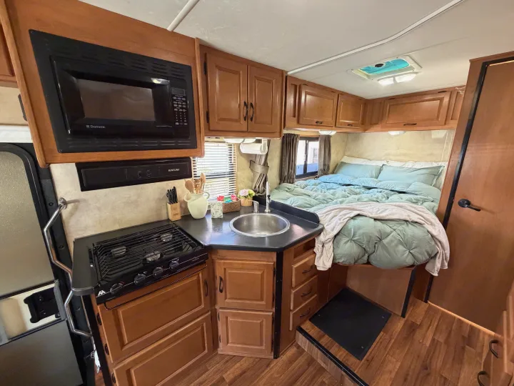 View of an RV rental interior: a compact galley kitchen with microwave, stove, and sink, alongside a comfortable bed.. Thor Majestic 2013