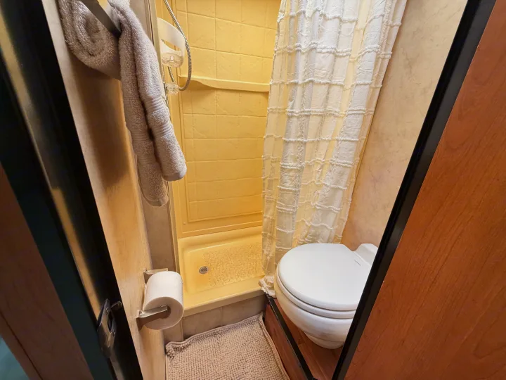 Compact RV rental bathroom with yellow shower, white toilet, ruffled curtain, towel, and toilet paper.. Thor Majestic 2013