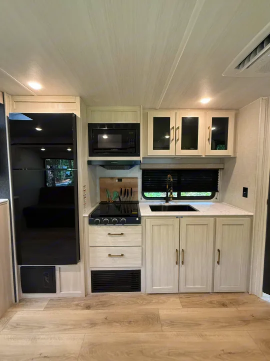 Modern RV rental kitchen featuring light wood cabinets, black appliances (fridge, microwave, stovetop), and a sink.. Forest River Surveyor Legend X 2025