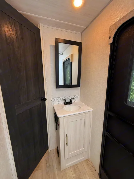 Functional RV rental bathroom with light vanity, dark faucet, sink, and mirror. Dark door and panel frame the compact space.. Forest River Surveyor Legend X 2025