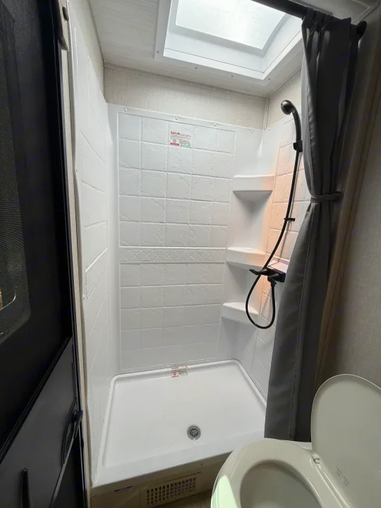 Inside an RV rental: a compact shower stall with shower head, grey curtain, skylight, and a partially visible toilet.. Forest River Surveyor Legend X 2025