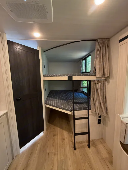 Cozy bunk beds with checkered mattresses in an rv rental. Features a black ladder, light wood floor, and a dark door.. Forest River Surveyor Legend X 2025