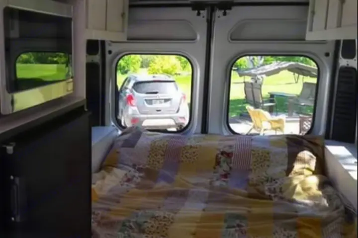 Interior of an rv rental: a bed, microwave & fridge visible. Rear doors frame outdoor views of a parked car and a patio setup.. RAM Dodge Promaster 2015