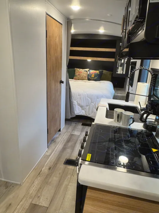 Inside an rv rental: a modern kitchen with a stovetop, sink, and coffee maker, leading to a cozy bedroom with a bed and pillows.. Keystone RV Springdale 2022