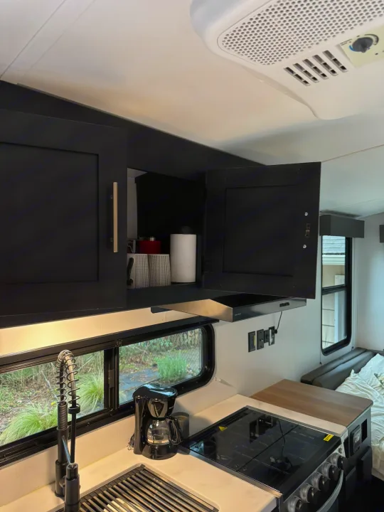 Inside an RV rental: a modern kitchen with black overhead cabinets (one open), sink, coffee maker, and stove. Compact travel living.. Keystone RV Springdale 2022