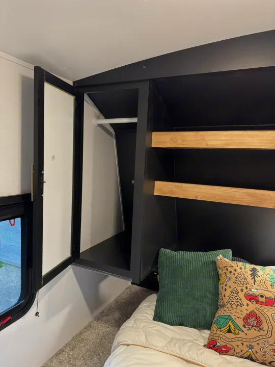 Cozy RV rental bedroom showing an open black wardrobe with hanging space and shelves above a bed with pillows.. Keystone RV Springdale 2022