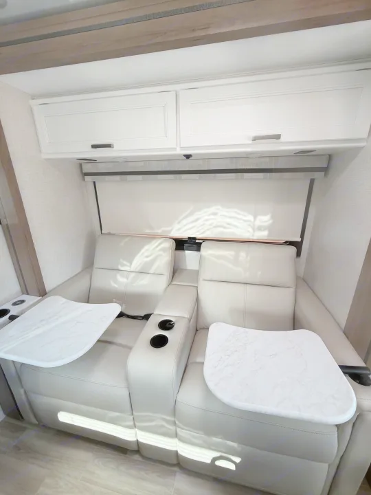 Inside an RV rental: two light gray recliners with cup holders, white marble-look tables, and overhead white cabinets.. Thor Vegas 2026