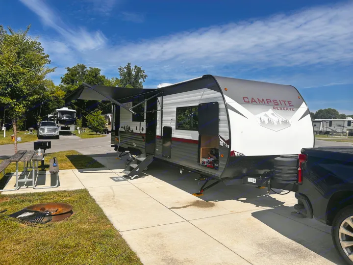 An RV rental travel trailer with awning extended and steps down, parked at a sunny campsite with picnic table and fire pit.. Forest River Campsite Reserve 2024