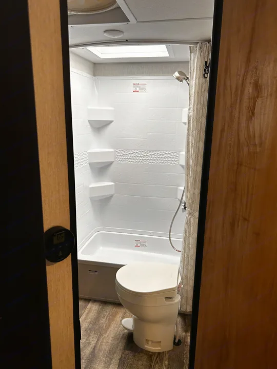 RV rental bathroom features a compact shower/tub combo, toilet, shelving, and wood-look floor. A thermostat is on the door frame.. Forest River Campsite Reserve 2024