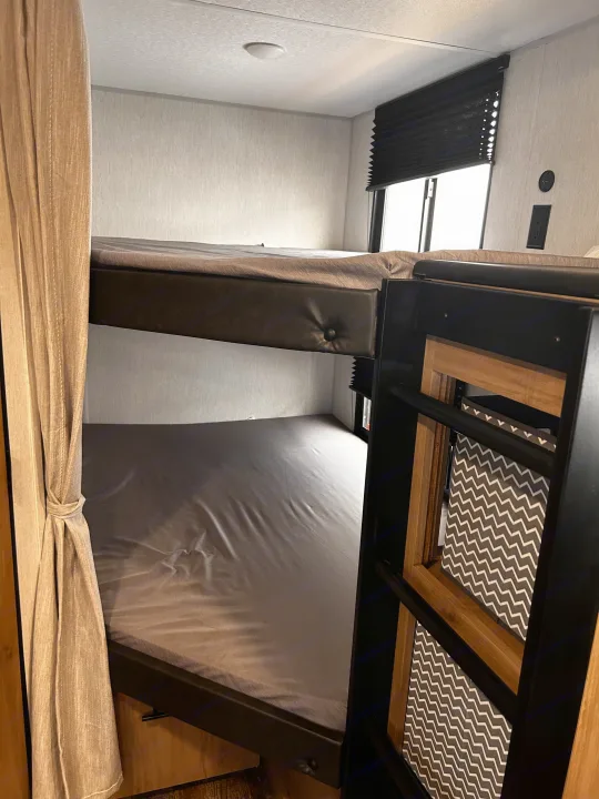 RV rental bunk bed setup: Two beds with gray sheets, privacy curtain, window with blind, and wall charging outlets.. Forest River Campsite Reserve 2024