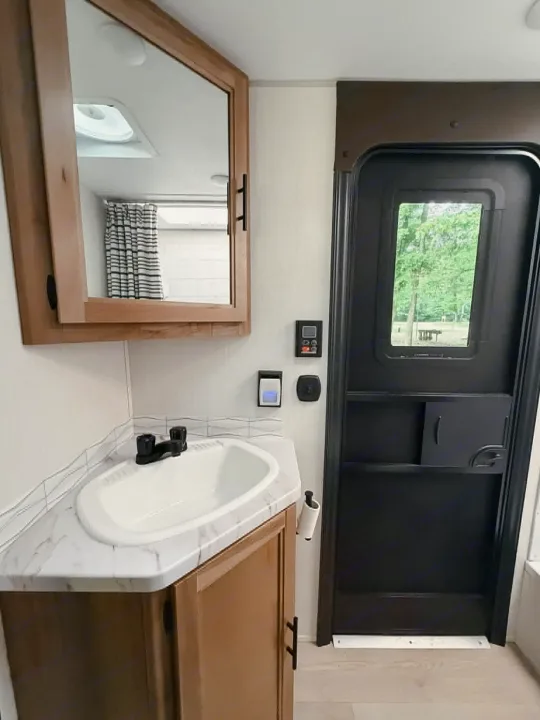 Inside an RV rental: a compact bathroom with a sink, mirrored cabinet, and an entry door viewing outdoor trees.. Forest River Cherokee Grey Wolf 2025