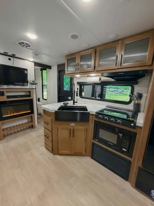 Modern RV rental interior featuring a full kitchen with dark sink, electric fireplace, TV, and light wood flooring.. Forest River Cherokee Grey Wolf 2025
