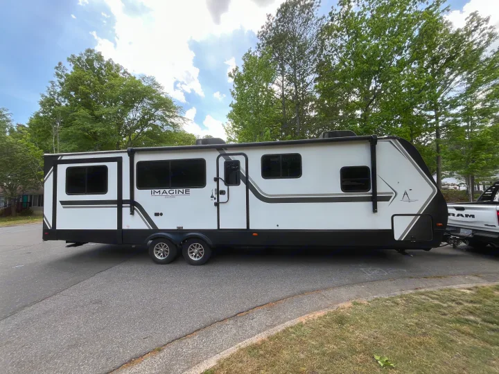 White Grand Design Imagine travel trailer RV rental parked on an asphalt road, hooked to a pickup truck under a partly cloudy sky.. Grand Design Imagine 3100RD 2025