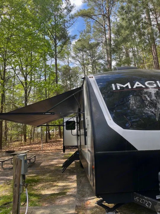 An Imagine Grand Design RV rental travel trailer at a wooded campsite with awning extended and utility hookups.. Grand Design Imagine 3100RD 2025