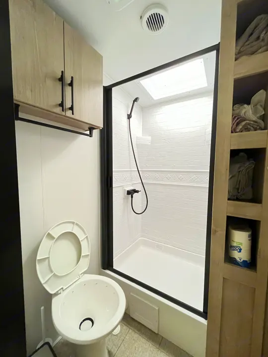 RV rental bathroom features a clean toilet, modern shower stall, overhead cabinets, and built-in shelving.. Grand Design Imagine 3100RD 2025