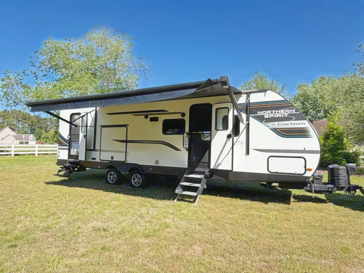 A white Northern Spirit by Coachmen travel trailer, an ideal RV rental, parked on green grass with its awning extended under a blue sky.. Coachmen 2963BH 2023