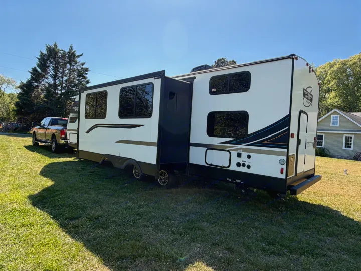RV rental: A white travel trailer with an extended slide-out is hitched to a pickup truck on a sunny lawn, near a house.. Coachmen 2963BH 2023