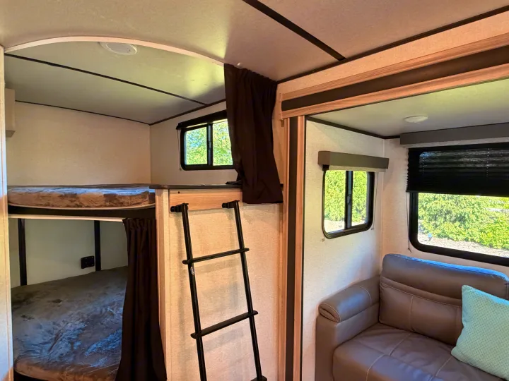 Interior of an RV rental: bunk beds with a ladder, grey sofa, and windows showing green trees outside.. Coachmen 2963BH 2023