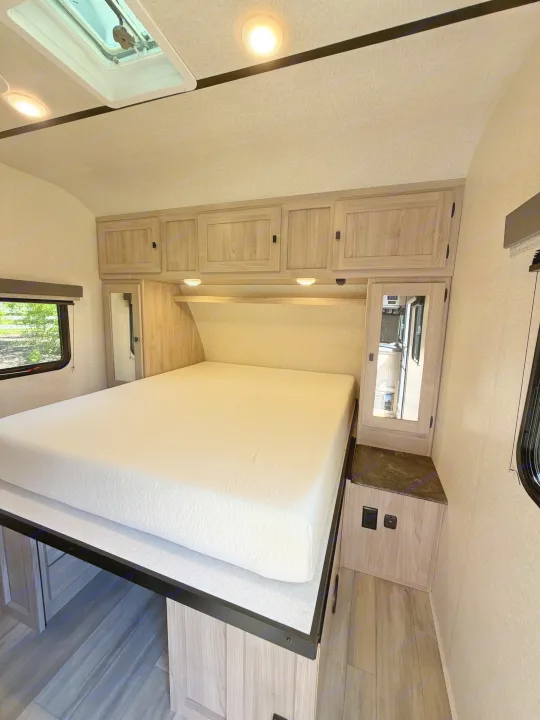 Bright RV rental bedroom features a large white bed, light wood cabinets, and windows with scenic views, perfect for an RV trip.. Coachmen 2963BH 2023