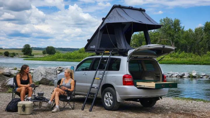 Two women enjoy a river view from their **SUV camping** setup with a **roof-top tent**. Ideal for your next **rv rental** **outdoor adventure**!. Toyota Highlander 2005