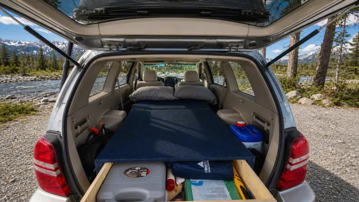 SUV car camping setup with a bed & storage, parked by a river with mountains. An alternative to an rv rental for outdoor adventures.. Toyota Highlander 2003