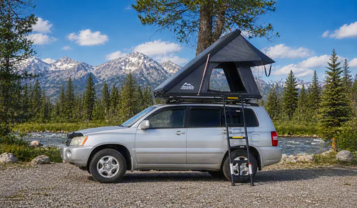 Silver SUV with a black rooftop tent and ladder, parked by a river with pine trees and snowy mountains. Great for an RV rental adventure.. Toyota Highlander 2003